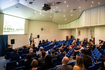 lecture theatre with people seated in the blue chairs listening to the speaker who is stood at the podium in front of them with a huge screen behind him